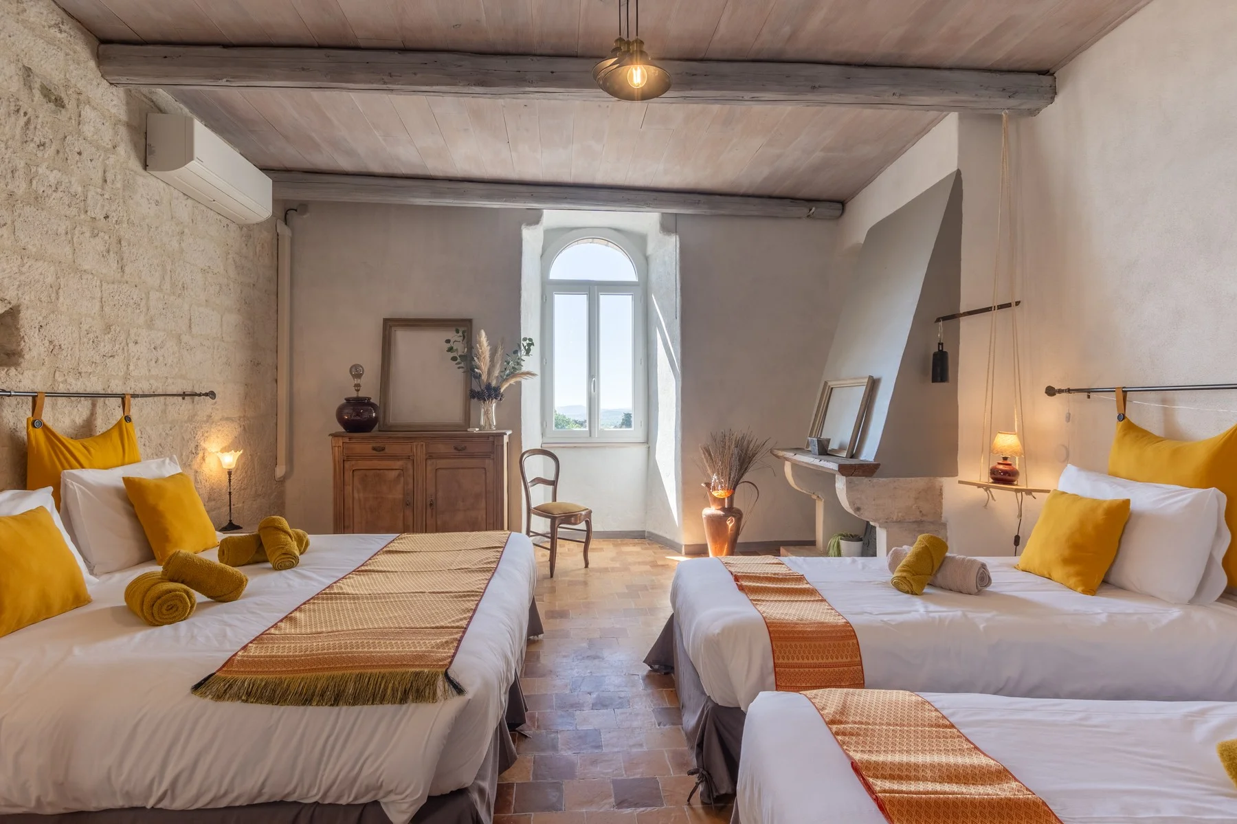 Chambre d'hôtel spacieuse en Ardèche avec trois lits, décoration jaune et pierre, lumière naturelle par baie vitrée