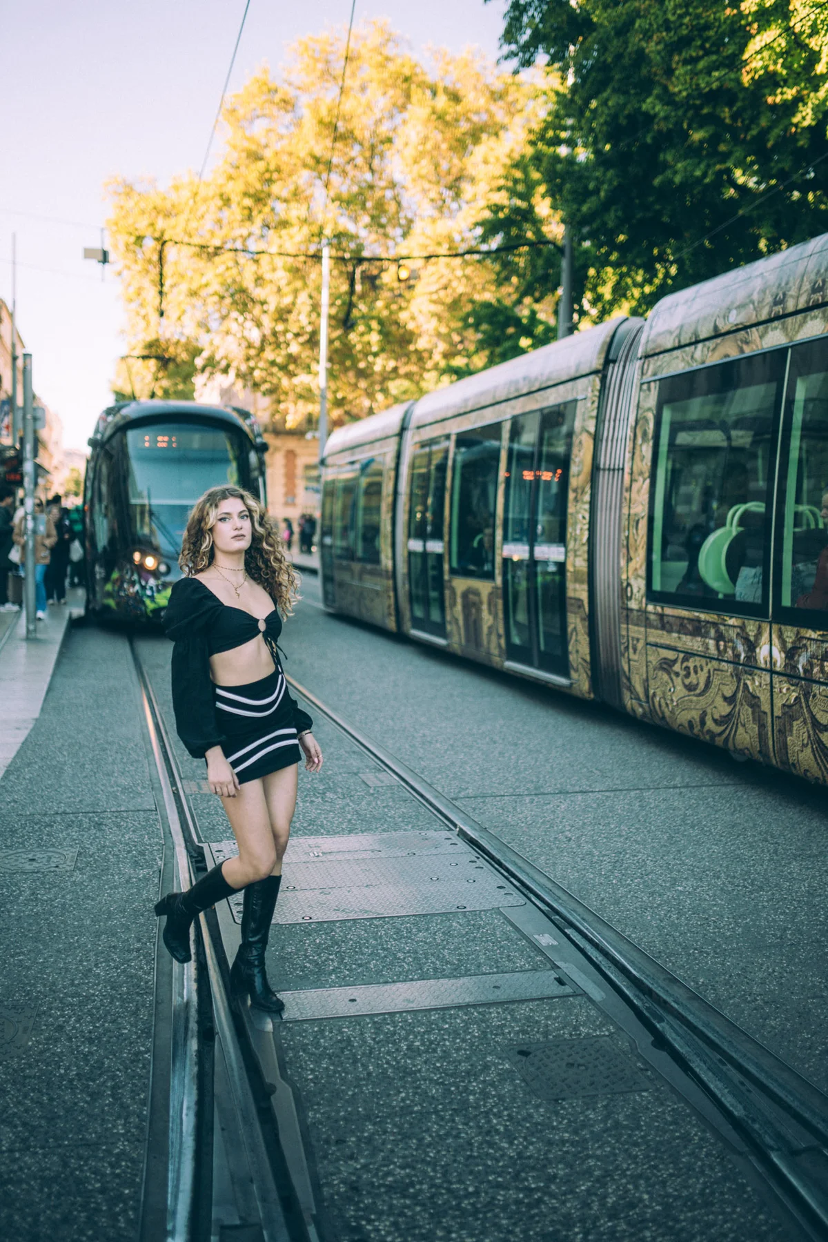 Shooting mode Montpellier, femme en tenue noire crop-top entre deux tramways, ambiance urbaine chic.
